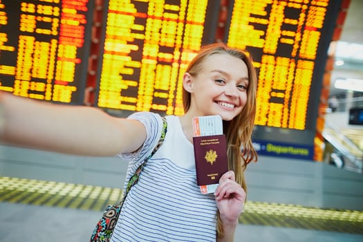 Beautiful young tourist girl in international airport, taking funny selfie with passport and boarding pass near flight information board
