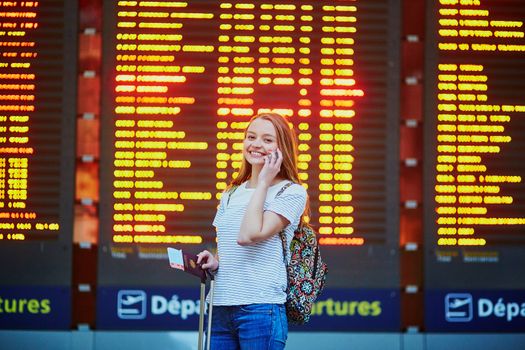 Beautiful young tourist girl with backpack and carry on luggage in international airport, near flight information board