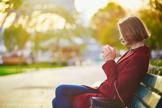 Beautiful young French woman sitting on the bench and drinking coffee near the Eiffel tower in Paris on a fall day