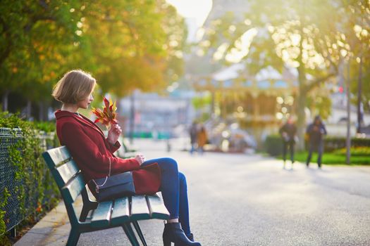 Beautiful young French woman with bunch of colorful autumn leaves sitting on the bench near the Eiffel tower in Paris on a fall day