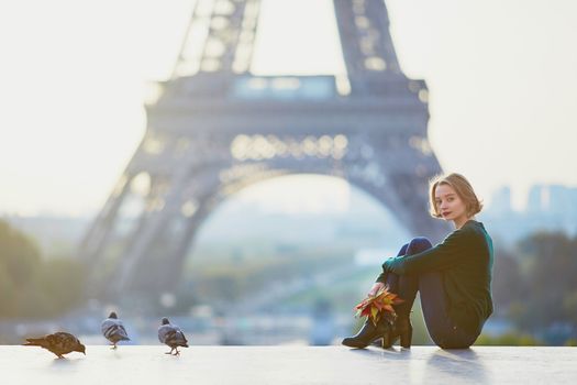 Beautiful young French woman with bunch of colorful autumn leaves near the Eiffel tower in Paris on a fall day