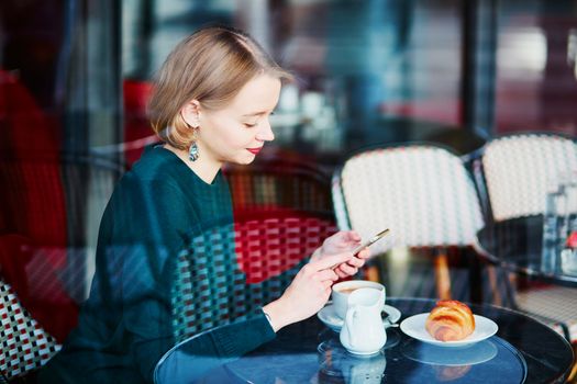 Young elegant woman drinking coffee and using her mobile phone in traditional cafe in Paris, France