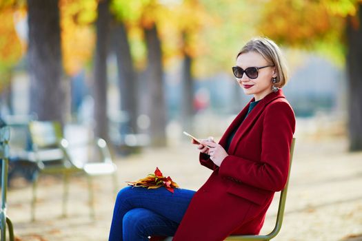 Elegant Parisian woman using her mobile phone in park on a bright sunny autumn day
