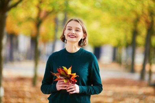 Beautiful young woman with bunch of colorful autumn leaves walking in park on a fall day. Luxembourg garden, Paris, France