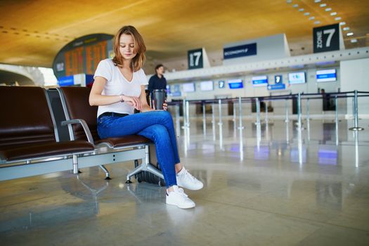 Young woman in international airport with luggage and coffee to go, waiting for her flight and looking at her watch