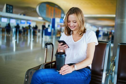 Young woman in international airport with luggage and coffee to go, waiting for her flight and looking at her phone