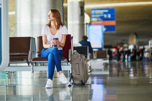 Young woman in international airport with luggage and coffee to go, waiting for her flight