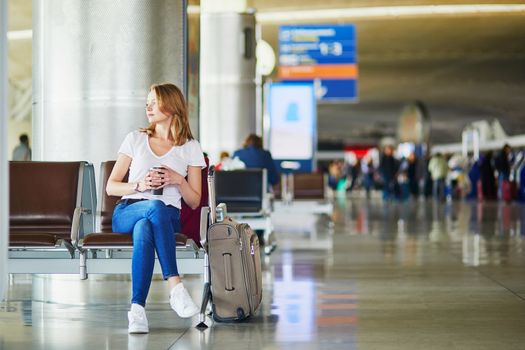 Young woman in international airport with luggage and coffee to go, waiting for her flight