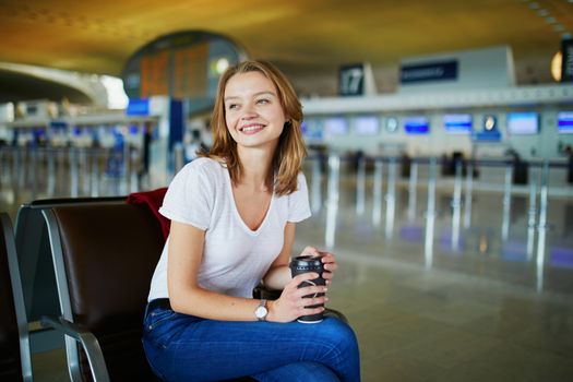 Young woman in international airport with luggage and coffee to go, waiting for her flight