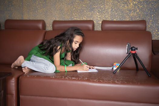 Concept of improper, lazy and way of studying during homeschooling or e-learning and make kids sleepy or boredom, young girl busy in writing by looking into mobile on sofa during covid-19 pandemic.