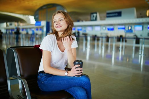 Young woman in international airport with luggage and coffee to go, waiting for her flight