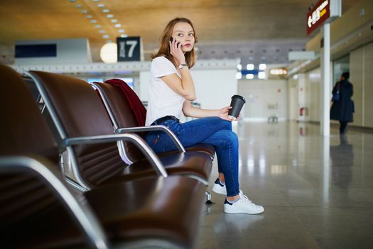 Young woman in international airport with luggage and coffee to go, waiting for her flight