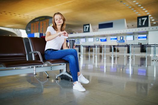 Young woman in international airport with luggage and coffee to go, waiting for her flight and looking at her watch
