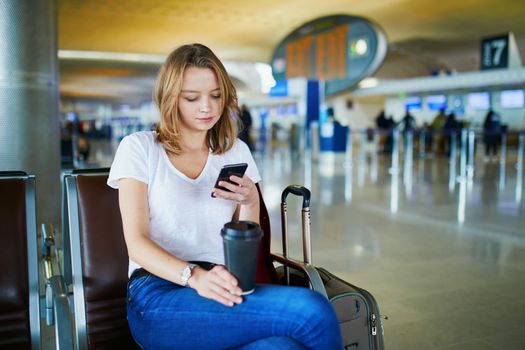 Young woman in international airport with luggage and coffee to go, waiting for her flight and looking at her phone