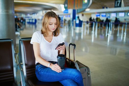 Young woman in international airport with luggage and coffee to go, waiting for her flight and looking at her phone
