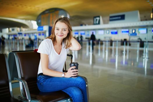 Young woman in international airport with luggage and coffee to go, waiting for her flight