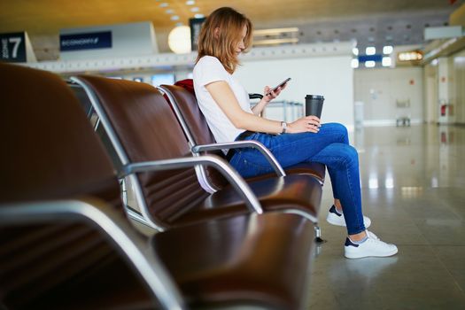 Young woman in international airport with luggage and coffee to go, waiting for her flight and looking at her phone