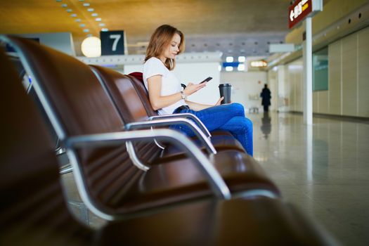Young woman in international airport with luggage and coffee to go, waiting for her flight and looking at her phone