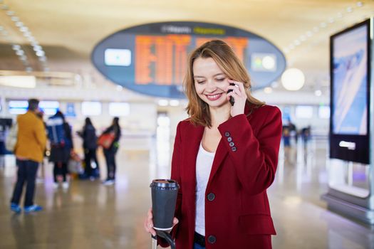 Young woman in international airport with luggage and coffee to go, speaking on the phone and waiting for her flight