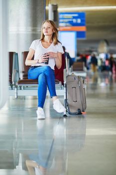 Young woman in international airport with luggage and coffee to go, waiting for her flight