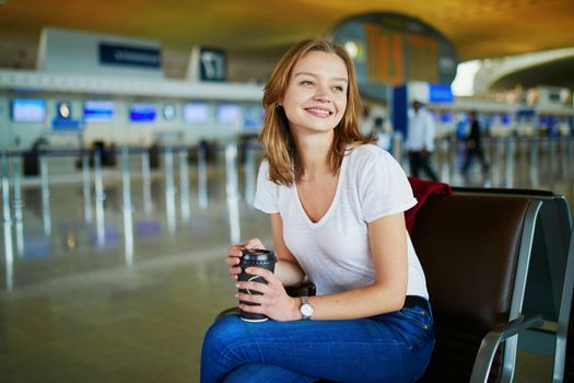 Young woman in international airport with luggage and coffee to go, waiting for her flight