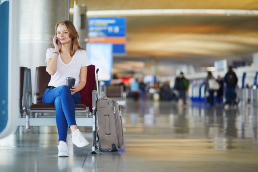 Young woman in international airport with luggage and coffee to go, waiting for her flight and speaking on the phone