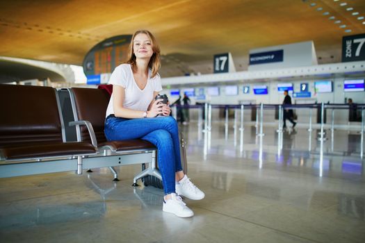 Young woman in international airport with luggage and coffee to go, waiting for her flight