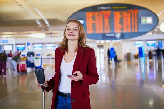 Young woman in international airport with luggage and coffee to go waiting for her flight