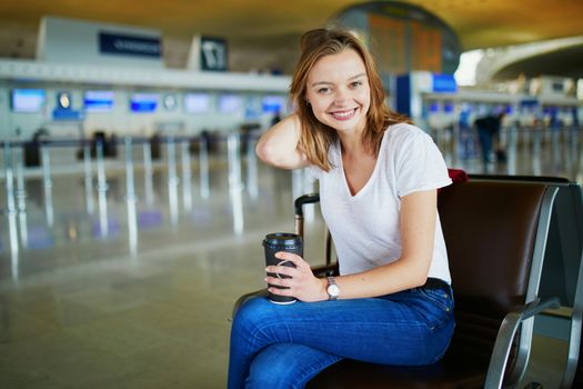 Young woman in international airport with luggage and coffee to go, waiting for her flight