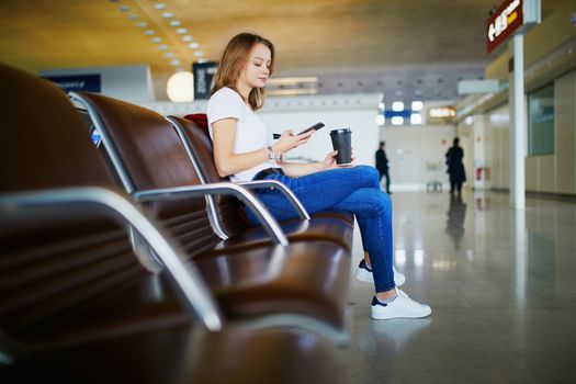 Young woman in international airport with luggage and coffee to go, waiting for her flight and looking at her phone