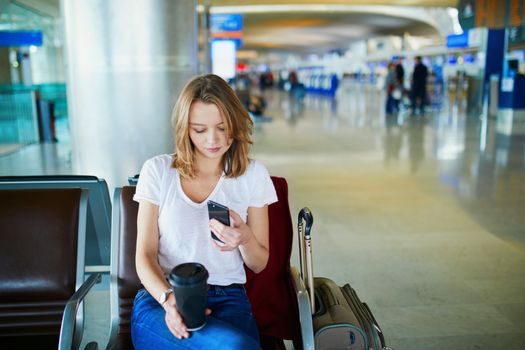 Young woman in international airport with luggage and coffee to go, waiting for her flight and looking at her phone