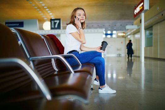 Young woman in international airport with luggage and coffee to go, waiting for her flight and speaking on the phone