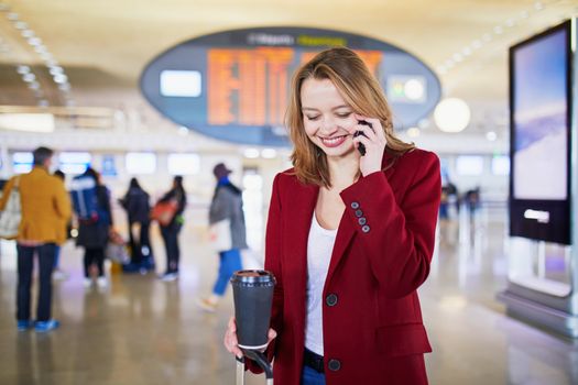 Young woman in international airport with luggage and coffee to go, speaking on the phone and waiting for her flight