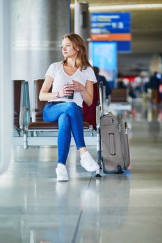 Young woman in international airport with luggage and coffee to go, waiting for her flight