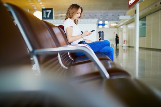 Young woman in international airport with luggage and coffee to go, waiting for her flight and looking at her phone