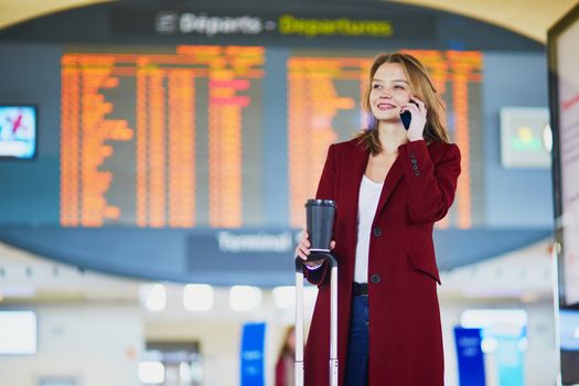 Young woman in international airport with luggage and coffee to go, speaking on the phone and waiting for her flight