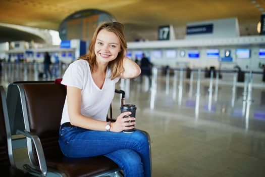 Young woman in international airport with luggage and coffee to go, waiting for her flight