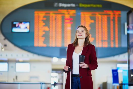 Young woman in international airport with luggage and coffee to go waiting for her flight