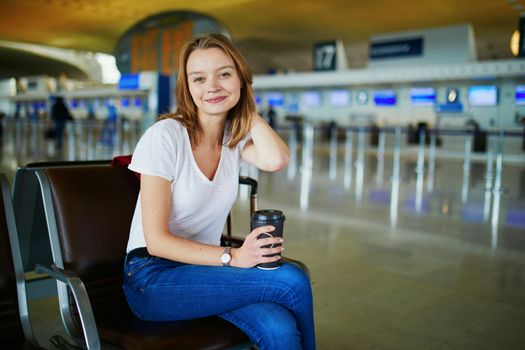 Young woman in international airport with luggage and coffee to go, waiting for her flight