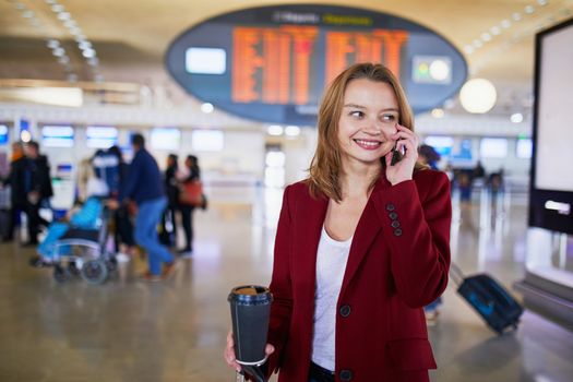 Young woman in international airport with luggage and coffee to go, speaking on the phone and waiting for her flight