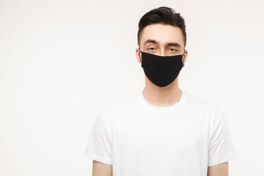 Young man in white t-shirt and black protective mask isolated on white background in studio. Healthy lifestyle concept, seasonal illness, and seasonal flu concept. Man wearing medical mask to prevent infection, airborne respiratory illness such as flu, 2019-nCoV.