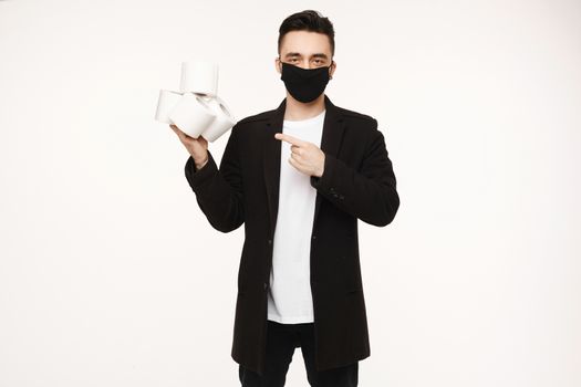 Handsome young man in black protective mask keeping four rolls of toilet paper and pointing finger on it, isolated on the white background. Young men in protective face mask represent a toilet paper.