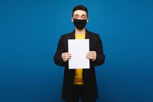 Young man in a black coat and black protective mask posing with an empty sheet of paper at the blue background, isolated with copy space. Promotion concept. Healthcare concept.