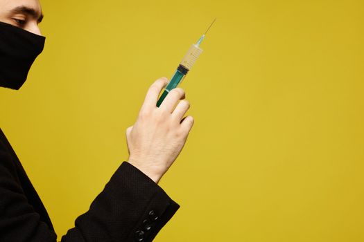 Side view of handsome man in black protective mask holding a syringe in his hand, isolated at the yellow background with copy space for your text. Concept of healthcare. Injection against viruses and flu