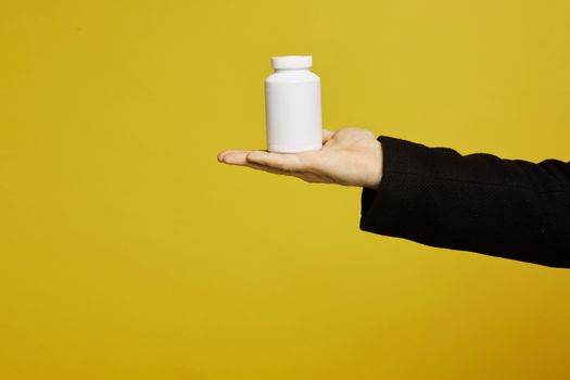A white bottle of drugs on a male palm on yellow background, isolated with copy space. Healthcare concept. Prevention infection, viruses and seasonal illness. COVID-19