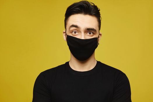 Handsome man in black t-shirt and black protective mask isolated on yellow background. Healthy lifestyle concept, seasonal illness, and seasonal flu concept. Young man wearing a protective mask to prevent infection and airborne respiratory illness