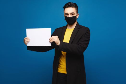Young shocked man in a black protective mask holding an empty sheet of paper and looking in the camera, isolated at the blue background. Promotion concept. Healthcare concept.