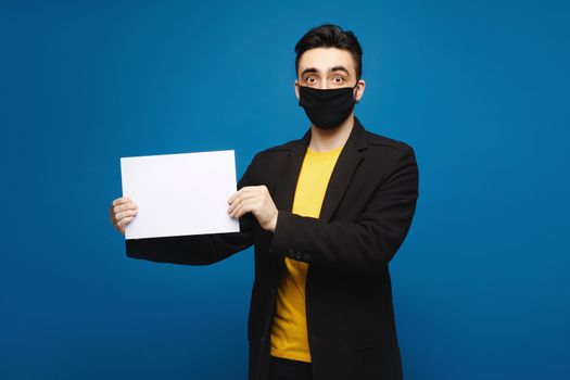 Young shocked man in a black protective mask holding an empty sheet of paper and looking in the camera, isolated at the blue background. Promotion concept. Healthcare concept.