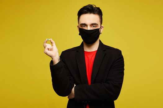 Handsome man in black coat and black protective mask keeps a pill in his hand and posing at the yellow background, isolated. Medicine for viruses. Healthcare lifestyle. Young man wearing a protective mask to prevent infection and airborne respiratory illness