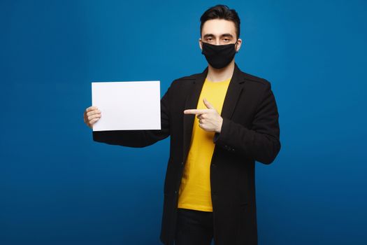 Young handsome man in black protective mask holding blank sheet of paper and pointing on it, isolated at the blue background. Promotion concept. Healthcare concept.